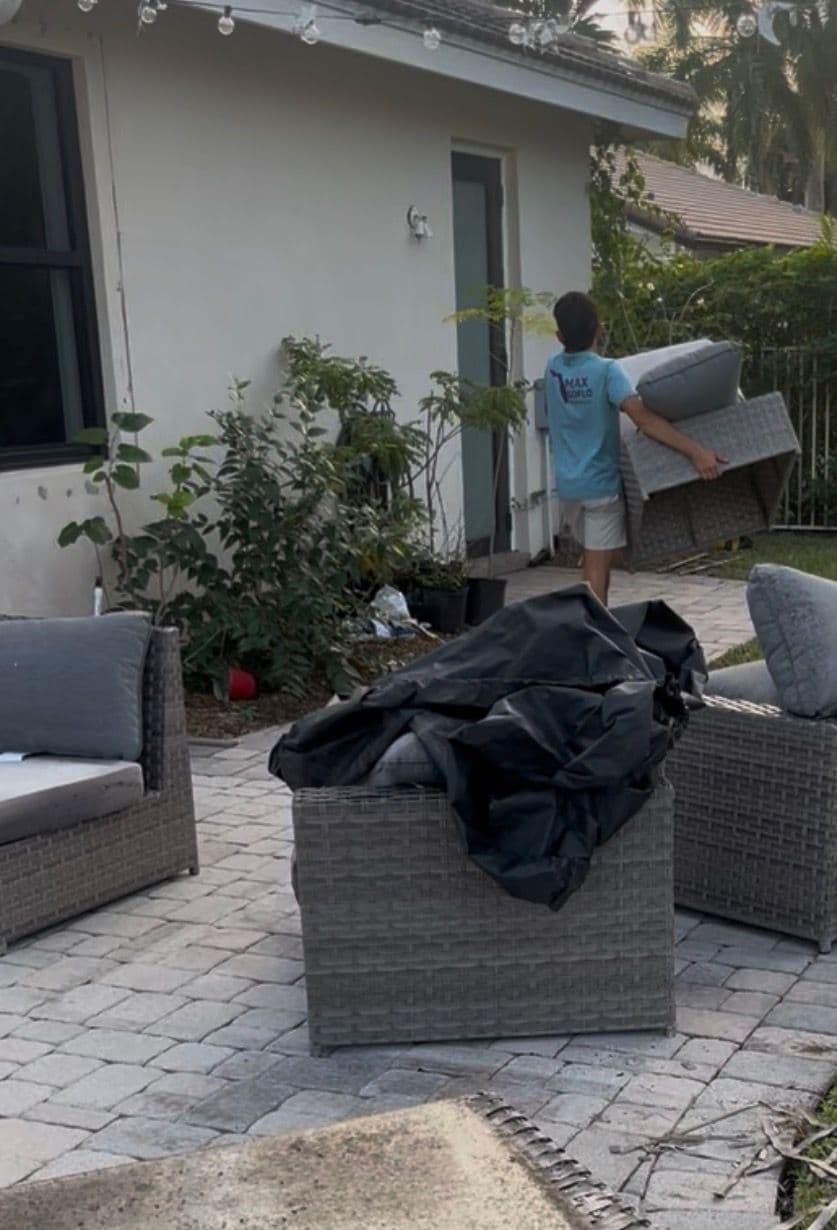 Person carrying outdoor furniture in a residential backyard with paver stones and plants.