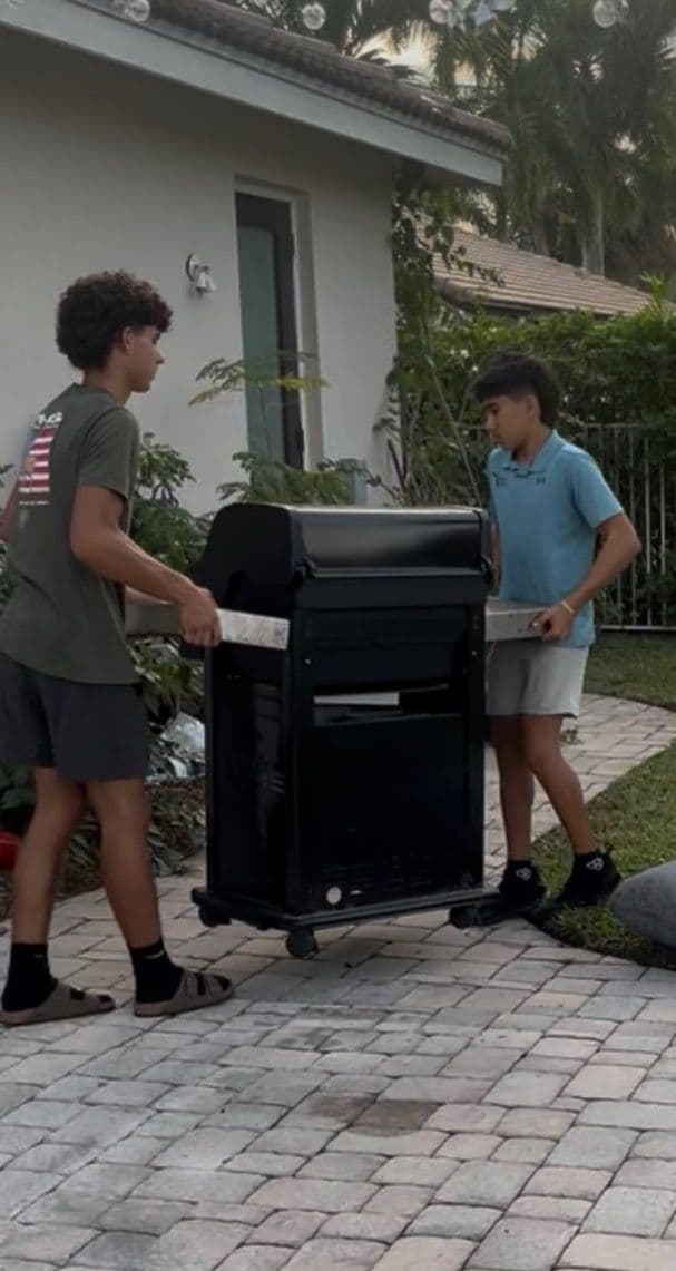 Two teenagers moving a large black grill outdoors on a residential patio.