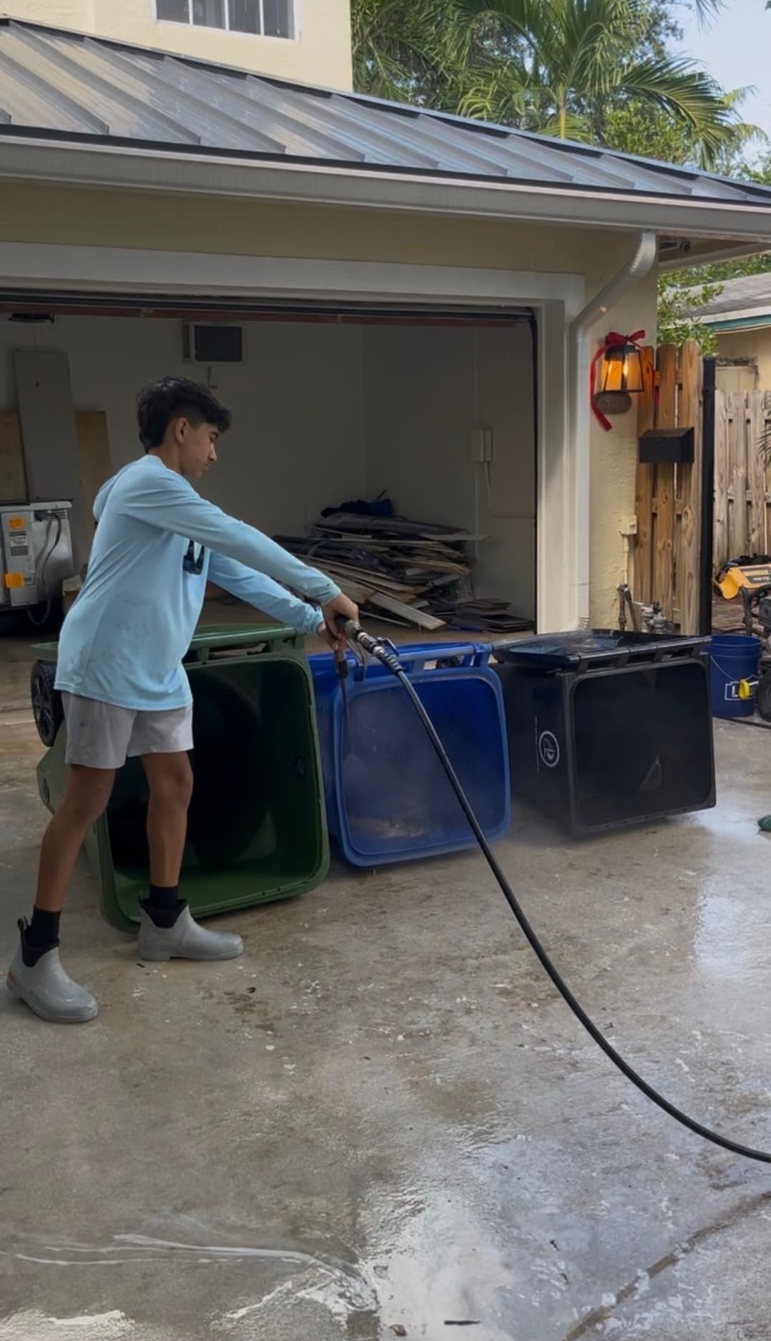 Boy washing garbage bins with a hose in a garage, surrounded by tools and equipment.
