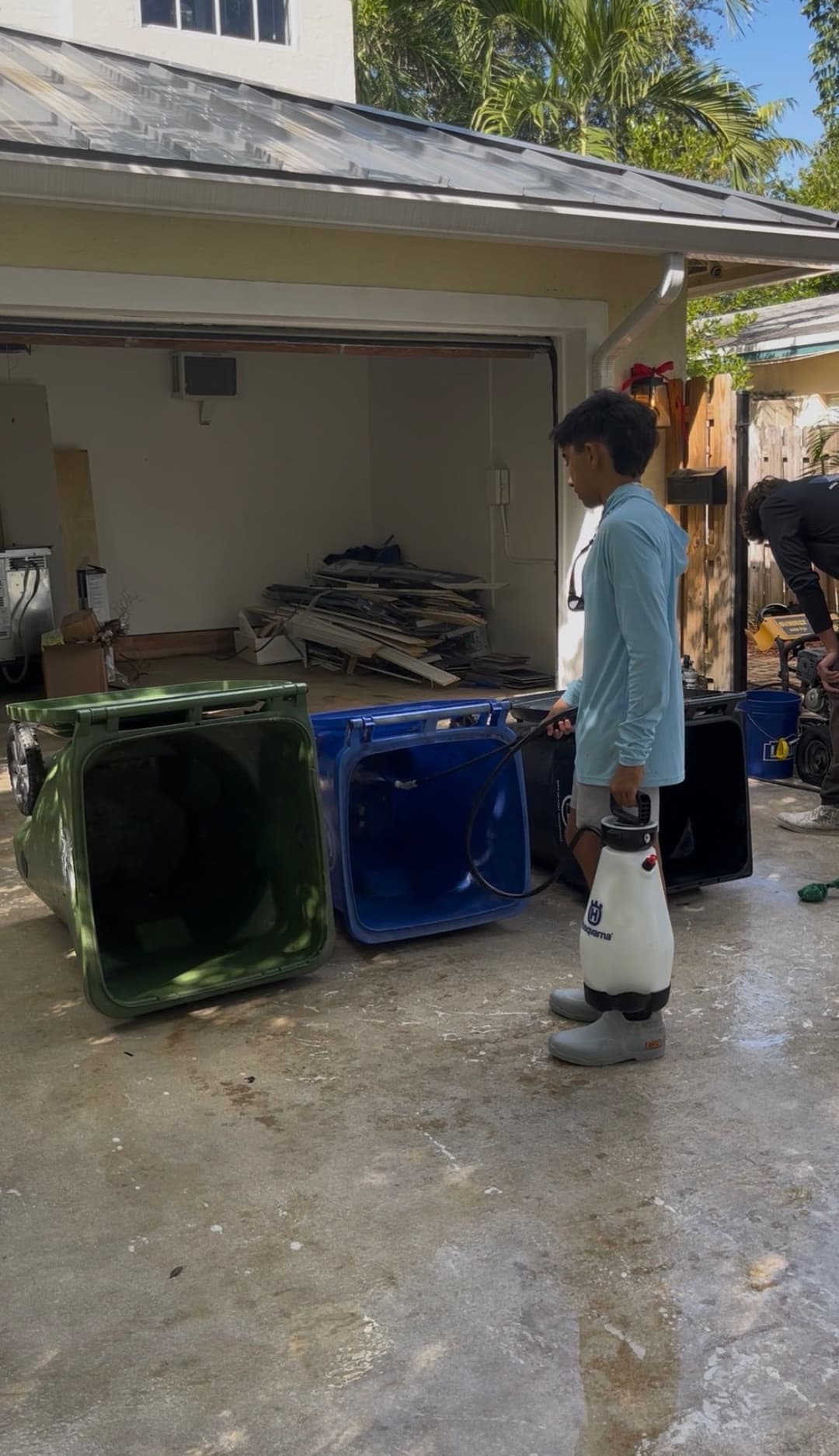 Child in boots cleaning recycling bins with a sprayer in a garage setting.