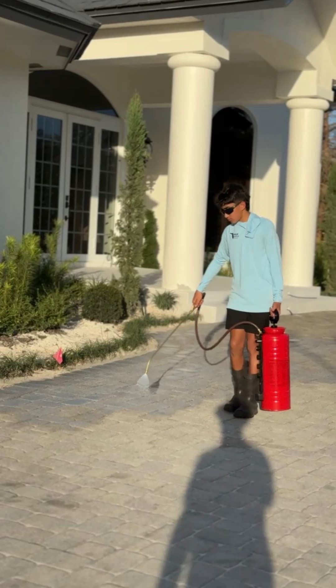 Child in blue shirt and sunglasses using a fire extinguisher on a driveway outside a home.