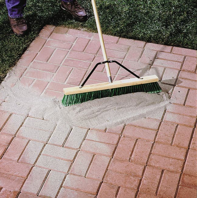 Person using a broom to sweep sand over brick pavers in a garden setting.