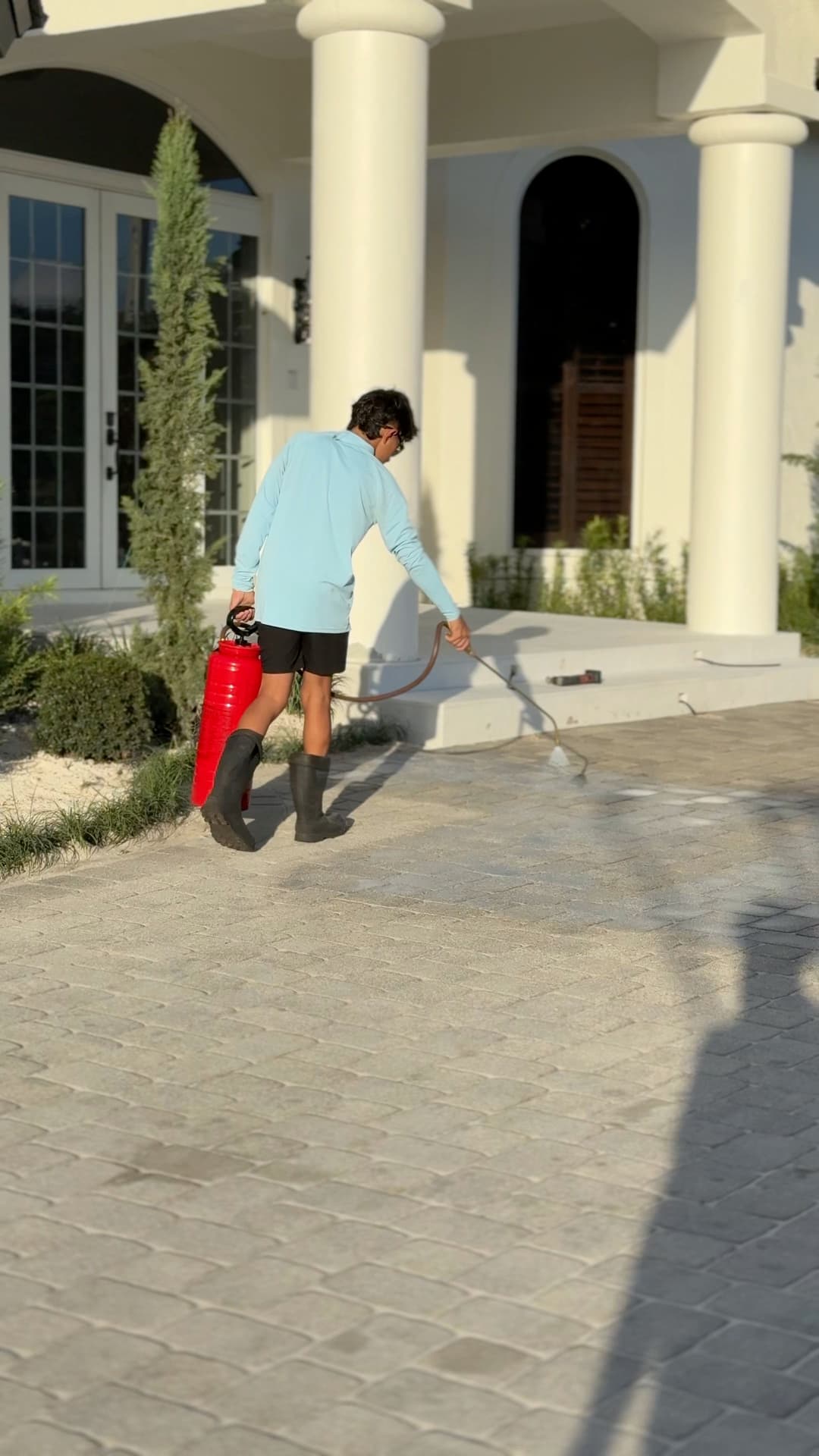 Person spraying a driveway with a red sprayer, wearing a light blue shirt and black boots.