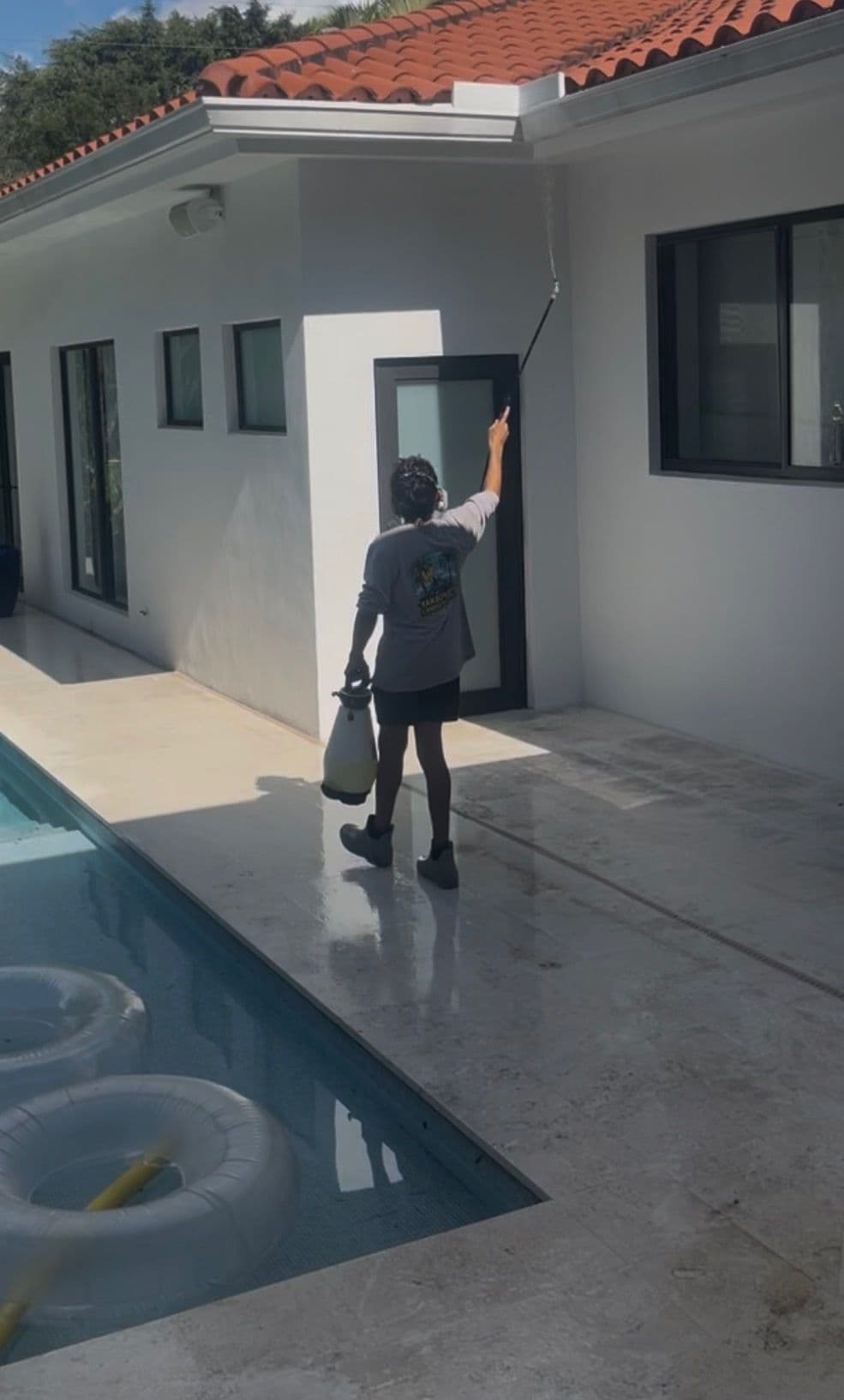 Person cleaning a house exterior with a sprayer near a pool on a sunny day.