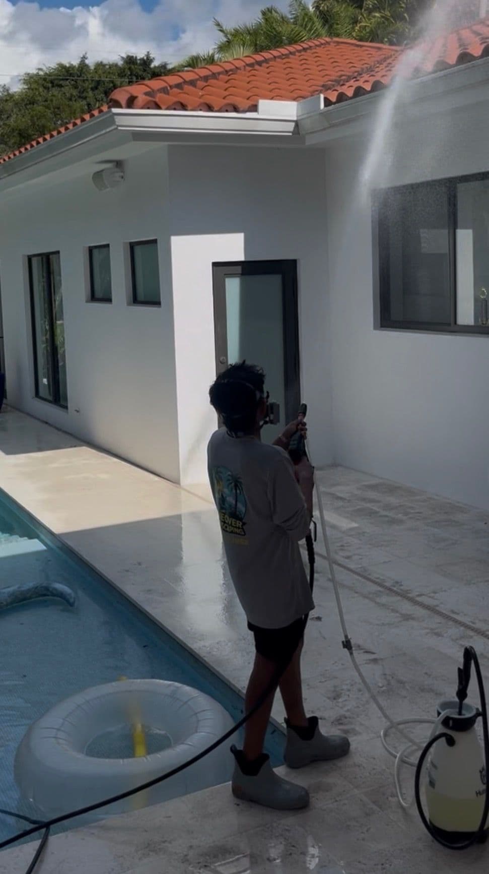 Person pressure washing a home's exterior near a swimming pool on a sunny day.