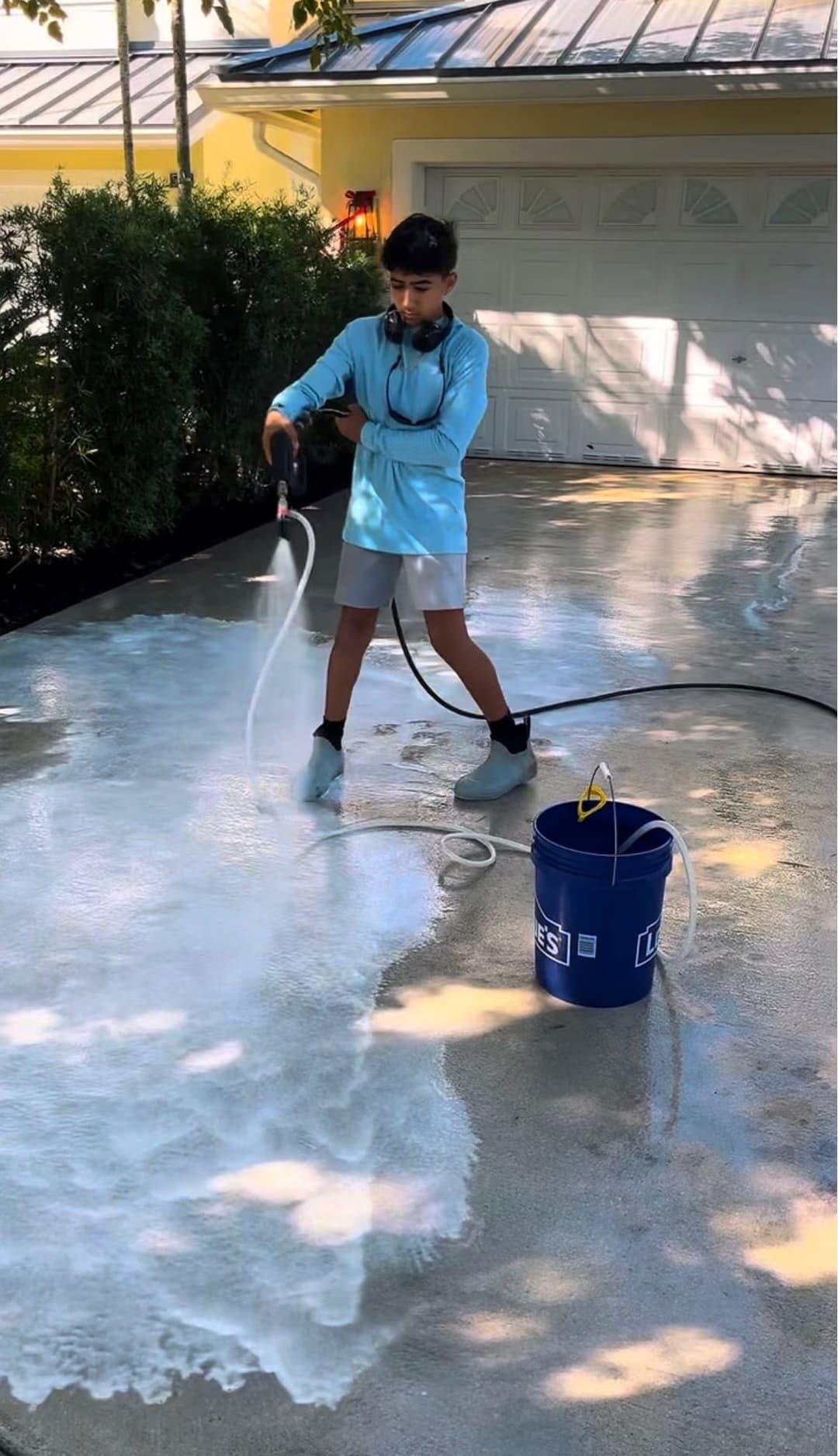 Child power washing a driveway, wearing gloves and using a hose with a bucket nearby.