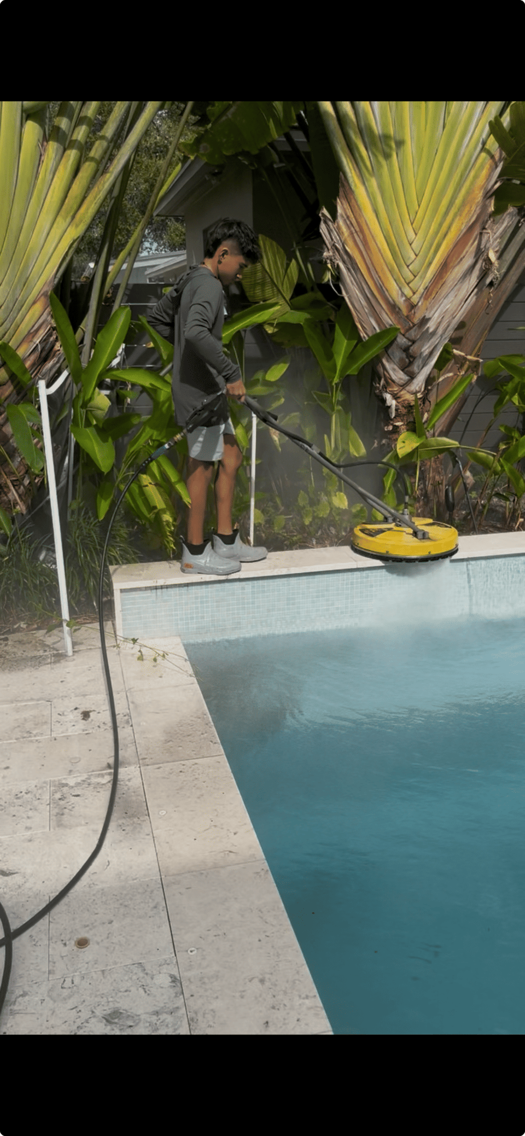 Person cleaning a pool with a pressure washer surrounded by lush tropical plants.