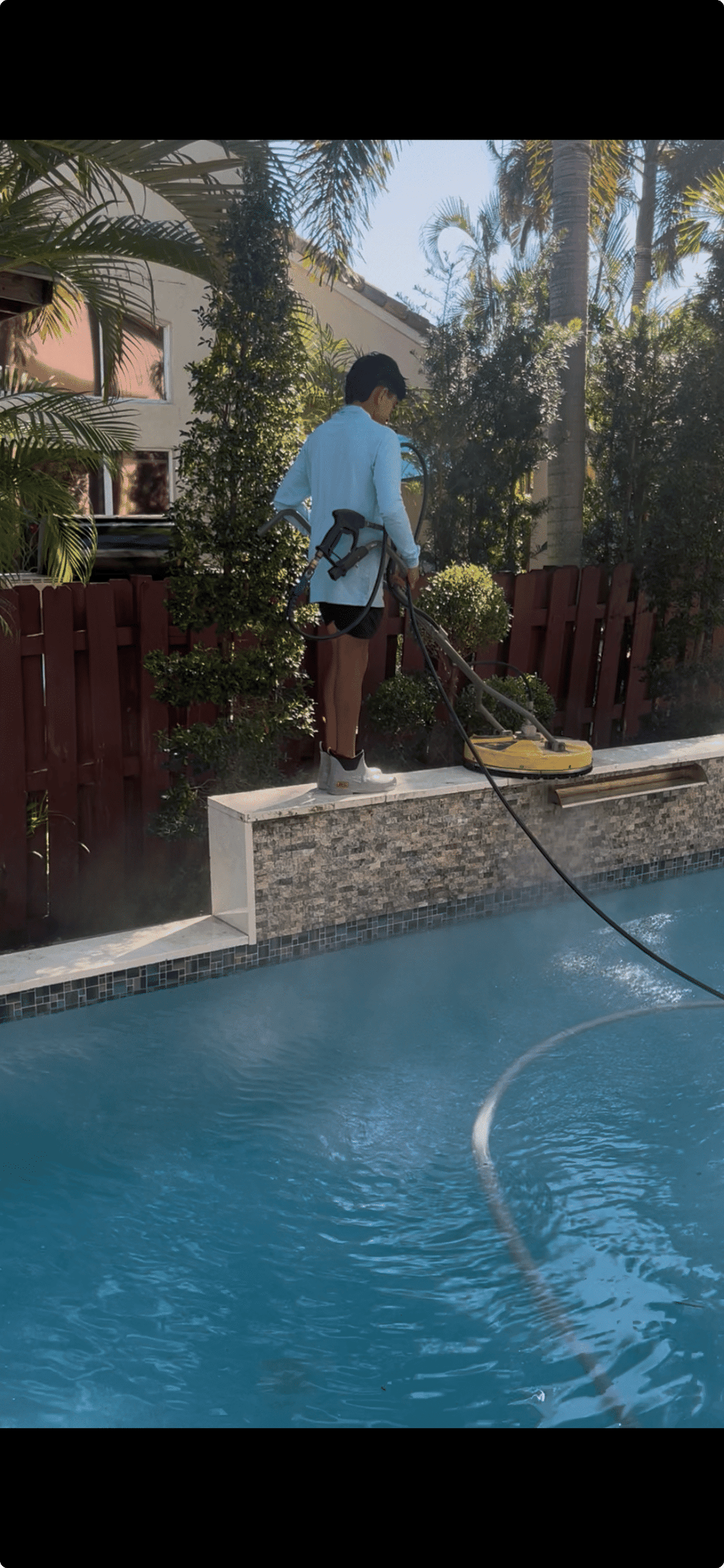 Person cleaning a pool with a pressure washer, surrounded by lush greenery and a wooden fence.
