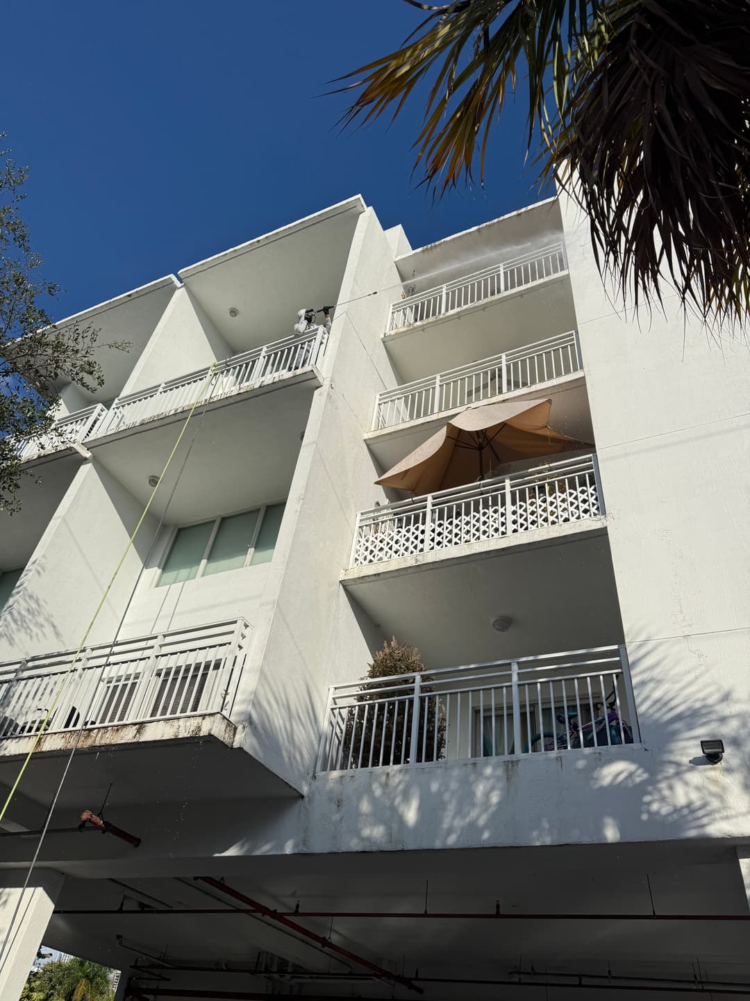 Four-story white apartment building with balconies and a patio umbrella against a blue sky.