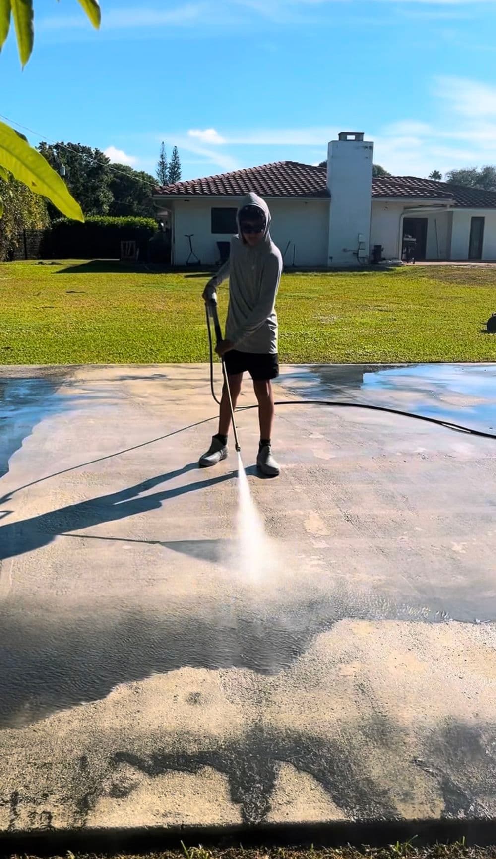 Person using a pressure washer on a driveway with a house and green lawn in the background.